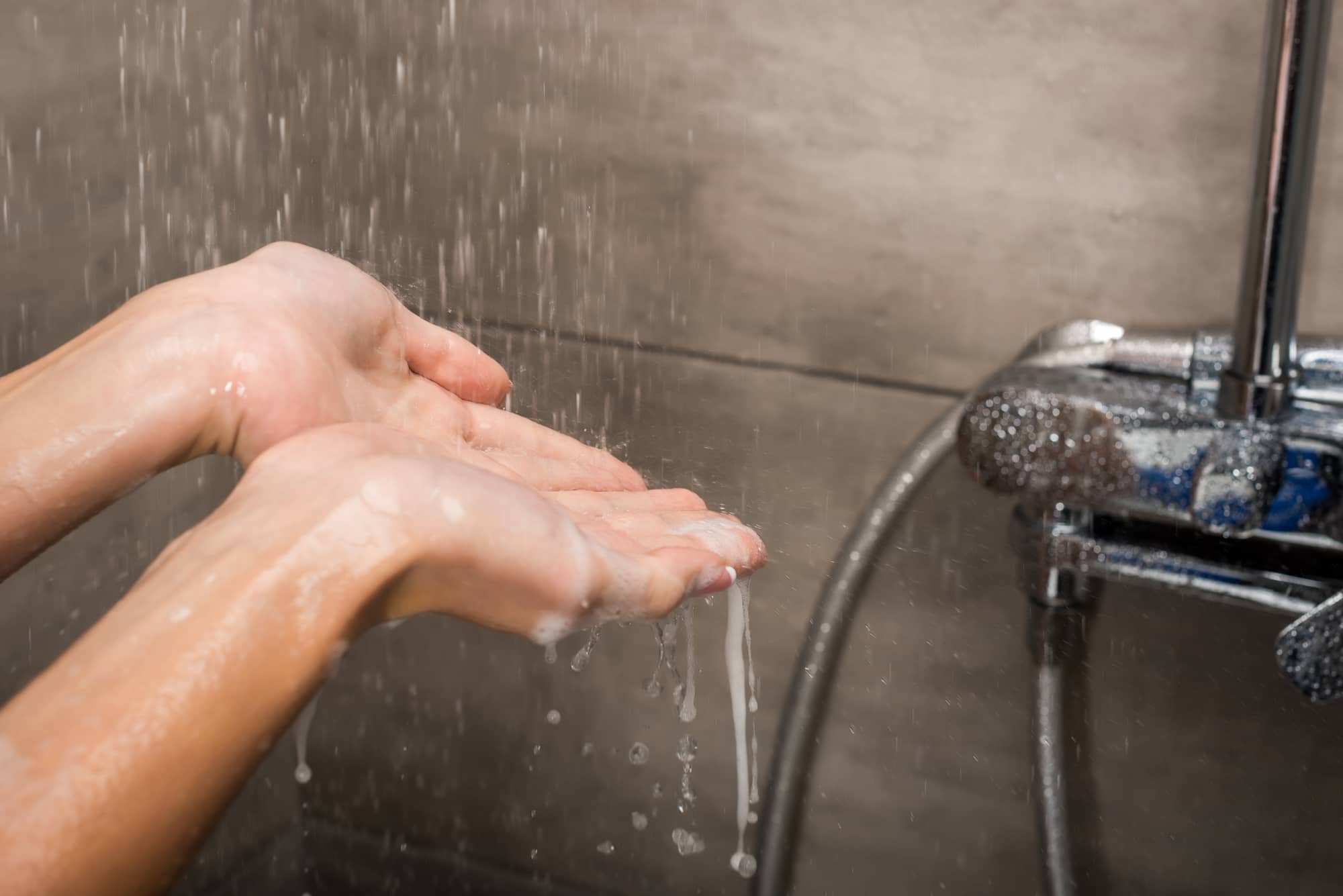 Eine Frau, die am Weg in die Dusche ist. Sie testet die Wassertemparatur unter der Wasserbrause. Im Hintergrund sind steingraue Fliesen und eine verchromte Wasserarmatur.